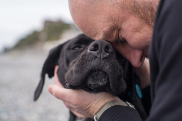 A man hugs a sad, black, long-eared dog on the beach by the sea on an autumn day, close-up, soft focus. Homeless street animals and people.