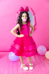 A young Hispanic girl with long curly hair wears a bright pink dress and a large pink bow. She blows a bubble with gum, standing in front of a pink background with balloons.