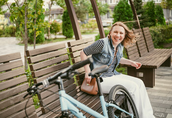 happy woman sits on a bench in park beside bicycle, celebrating a sunny day with leisure activities and a cheerful smile