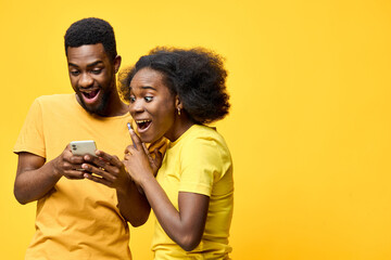 Happy young African American couple sharing exciting news on smartphone against a vibrant yellow background, expressing joy and surprise