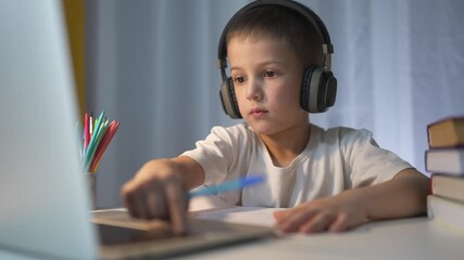 Young boy with headphones and laptop. Schoolboy is learning how to use laptop. A lifestyle child studying on a table at evening. A young child using headphones and a laptop.