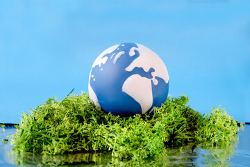 A blue and white globe is placed on vibrant green moss, symbolizing Earth Day. The backdrop is a bright blue color, enhancing the theme of environmental awareness and nature