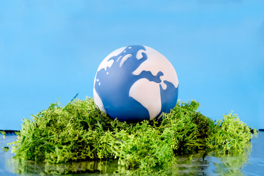 A blue and white globe is placed on vibrant green moss, symbolizing Earth Day. The backdrop is a bright blue color, enhancing the theme of environmental awareness and nature