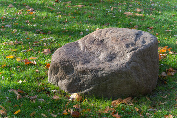 A weathered boulder rests on green grass dotted with fallen leaves. Soft afternoon light shapes the stone. © Jakob