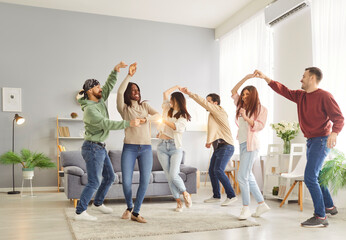 Naklejka premium Group of cheerful friends dancing in pairs to energetic music and having fun together in living room. Happy multinational buddies enjoying weekend party and celebrating special occasion at home.