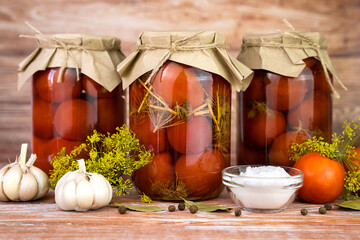 Pickled homemade tomatoes with garlic, dill, and spices in jars on the wooden table at the kitchen. Autumn food storage. Close-up.