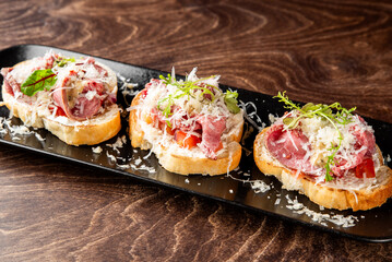 Three gourmet bruschetta appetizers with roast beef, parmesan cheese, and microgreens on a black plate against a dark wood background. Perfect for restaurant menus and food blogs.