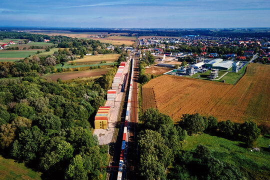 Aerial view of freight train loaded with shipping containers on railway station in small town near fields and industrial buildings. Concept of transportation, logistics and infrastructure