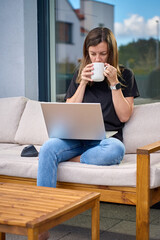Woman sitting on sofa using laptop with wireless mouse and coffee cup. Person relaxing on home patio with wooden outdoor furniture. Concept of remote work, comfort and modern home lifestyle