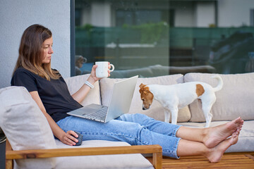 Woman and her dog sitting on terrace sofa with laptop and coffee cup. Concept of remote work, relaxation and life with pet at home