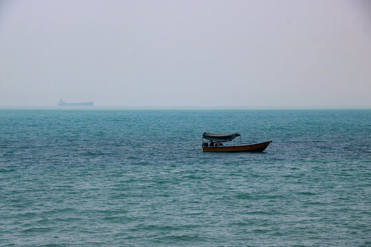 Small Fishing Boat Drifting on Choppy Turquoise Sea with Distant Cargo Ship in Heavy Fog
A wide shot capturing a sense of isolation and scale. A small, yellow boat, possibly a fishing vessel, sits alo