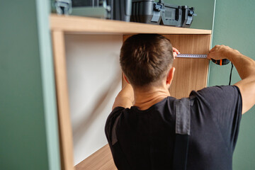 Man measuring wooden cabinet, using measuring tape and pencil to mark panel during home furniture assembly