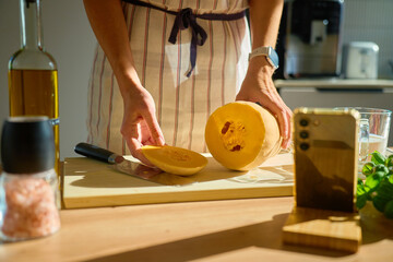 Person in apron holding sliced butternut squash on cutting board in kitchen. Woman slicing pumpkin on table with olive oil, spices, vegetables and smartphone. Concept of cooking and healthy food