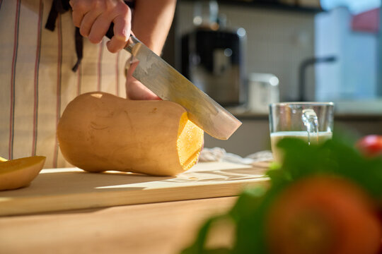Person in apron cutting butternut squash on wooden board in kitchen. Close-up of woman slicing fresh pumpkin with fresh vegetables and herbs on table. Concept of healthy eating and home cooking