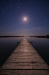 Serene wooden pier under a vast, star-filled night sky with a luminous moon guiding the way across tranquil waters