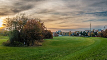 Jesienny krajobraz miejskiego parku o zachodzie słońca. Po lewej stronie znajduje się kępka drzew z brązowymi liśćmi i gołymi gałęziami. Niebo jest dramatyczne, z szarymi, rozciągniętymi chmurami 