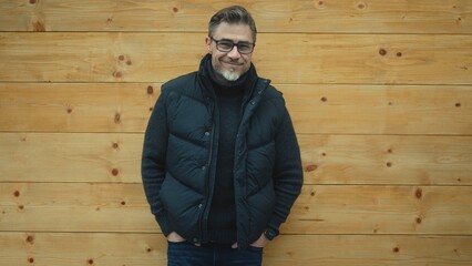 Portrait of smiling man wearing glasses, sweater and winter vest, standing against wooden wall with...