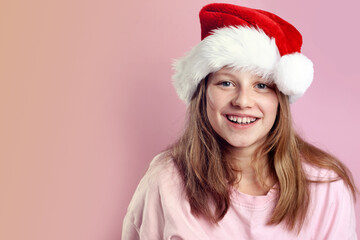Excited pretty 11-year-old girl in pink T-shirt and Santa hat posing on pink background