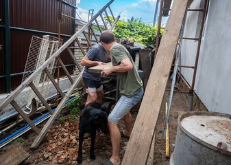 Two men carry a wooden plank amidst construction debris in a cluttered backyard, accompanied by a black dog. Shows teamwork and hard work during renovation or cleanup projects outdoors.