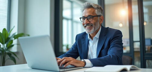 Happy senior businessman works on modern laptop in bright office. Indian man types on keyboard, looks satisfied, successful. Mature entrepreneur manages finances, online trading, planning market