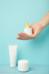 Set of various cosmetic products in blank packaging, creams in jars, serum in bottle on blue background. Woman holding bottle of serum