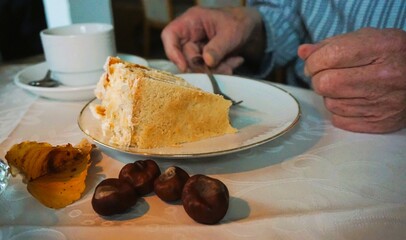 Alter Mann sitzt am Tisch und isst mit Kuchengabel frische Stachelbeertorte vom Teller auf Tisch mit weißer Tischdecke und braunen Kastanien in Café am Nachmittag im Herbst