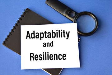 ADAPTABILITY and RESILIENCE - words on a white sheet on a black notebook with a magnifying glass