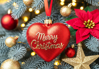 Close-up of a red heart-shaped "Merry Christmas" ornament hanging on a blue-toned pine tree, surrounded by gold ornaments, lights, and a poinsettia.