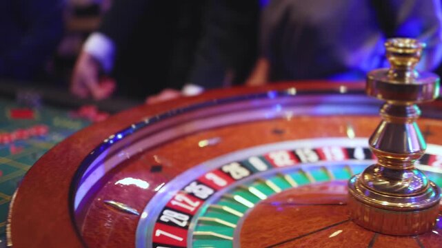 Casino table with roulette wheel in motion, with casino chips, tokens, the hand of croupier, dollar money and a group of gambling rich people playing bet on a party, blue poker table and deck of cards