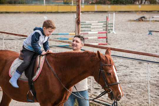 Happy father walking next to his young son riding a brown horse on a bridle path in an equestrian training center on an autumn day