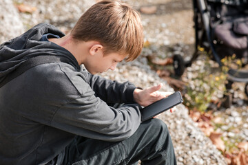 Young teenage boy sitting outdoors, focused on using a black smartphone with a blank screen on a sunny day