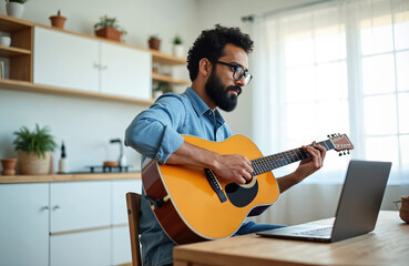 Indian musician sits at desk analyzing laptop screen playing acoustic guitar in bright dining room. Young composer creates music using digital tools, traditional instrument. Focuses on songwriting
