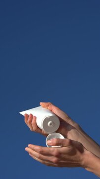 Female hands holding white mockup tube, opening it and applying cream on palm of their hand against clear blue sky. Concept of daily face cream for natural beauty, hydration, and lifestyle skincare
