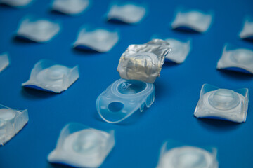 Side view in focus is an open container with contact lenses on top of which lies a transparent blue lens against a background of contact lens containers on a blue background. Concept of vision