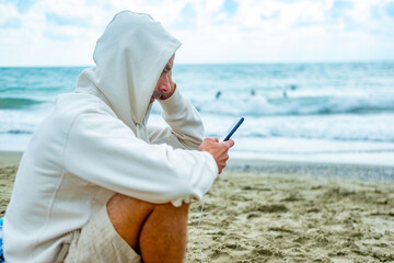 young Man in white hoodie sits on sandy beach, hood up, focused on smartphone, ocean waves behind him, capturing modern solitude, hooded figure modern isolation, digital escape, and coastal disconnect