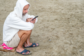 young Man in white hoodie sits on sandy beach, hood up, focused on smartphone, ocean waves behind him, capturing modern solitude, hooded figure modern isolation, digital escape, and coastal disconnect