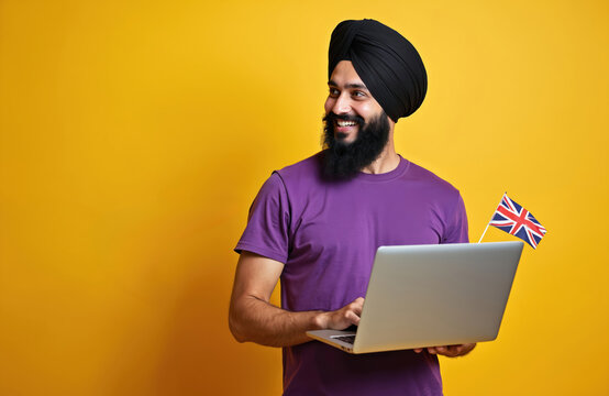 Smiling sikh man works on laptop holds British flag. Person study english language. specialist work remotely on yellow studio background. Indian man smiles wears turban, casual clothes using modern