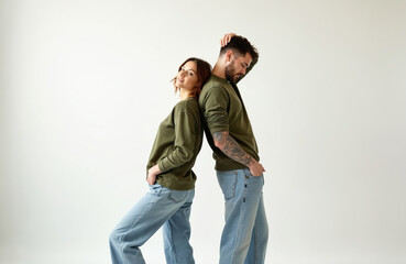 Young couple standing back to back wearing matching green sweaters and jeans. Woman smiles at camera, man looks down with hand on head. Casual fashion studio shoot.