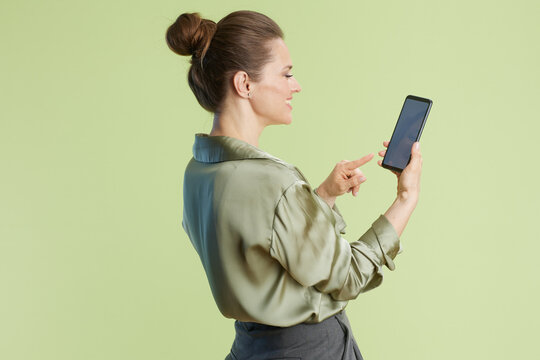 A woman smiles and touches the screen of a black smartphone. The profile view, green blouse, and grey pants suggest a modern, professional lifestyle.