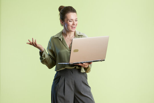 A middle-aged woman smiles while presenting on her laptop, gesturing with her hand. She wears a green blouse and gray pants against a bright, light green studio backdrop. - Powered by Adobe