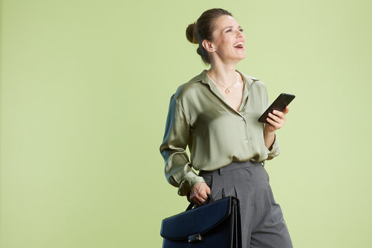 Smiling woman with a smartphone and briefcase, looking upwards with a joyful expression.  Light green blouse and gray pants against a soft green backdrop suggest success.