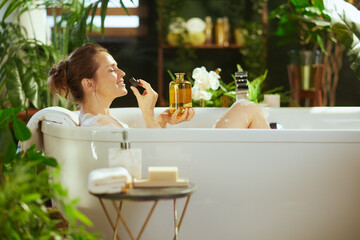 A smiling woman with closed eyes relaxes in a white bubble bath, holding essential oil bottles for aromatherapy. Lush plants & bath products create a tranquil, luxurious home spa for deep self-care.