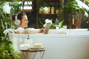 An adult woman in a bubble bath beams, enjoying her digital tablet & headphones amidst a lush, green bathroom. Natural light & a serene atmosphere underscore a perfect moment of self-care & relaxation
