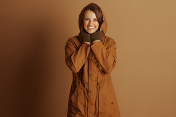 A woman with brown hair smiles warmly, hands clasped at her chin, dressed in a brown hooded parka...