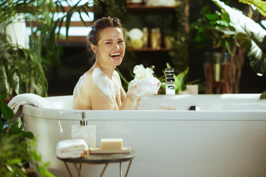 A woman with a bun hairstyle laughs joyfully in a white bubble bath, holding foam. Surrounded by lush green plants in a spa-like bathroom, captures pure happiness & relaxed self-care.
