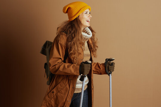 A smiling young woman, outfitted in a yellow beanie, brown coat, and patterned sweater, holds trekking poles and wears a backpack against a warm studio background, exuding adventure.