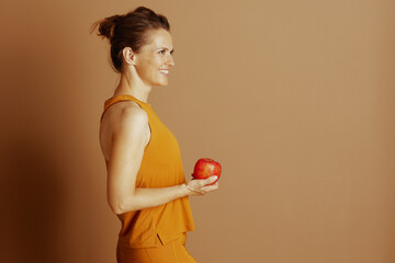 A fair-skinned woman in bright orange activewear smiles, holding a red apple in profile against a...