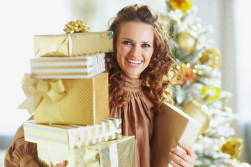 A smiling woman with curly brown hair, wearing a brown turtleneck, holds a tall stack of gold-wrapped Christmas presents, set against a softly lit, decorated Christmas tree, conveying holiday joy.