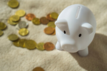 white ceramic piggy bank standing on a beige towel, euro coins scattered in the background with a narrow depth of field, concept of finance, saving money, investment.