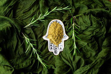 Organic henna powder with rosemary sprigs in a ceramic hamsa dish on green silk fabric. Natural herbal dye for body art, hair coloring, and Ayurvedic beauty care.
